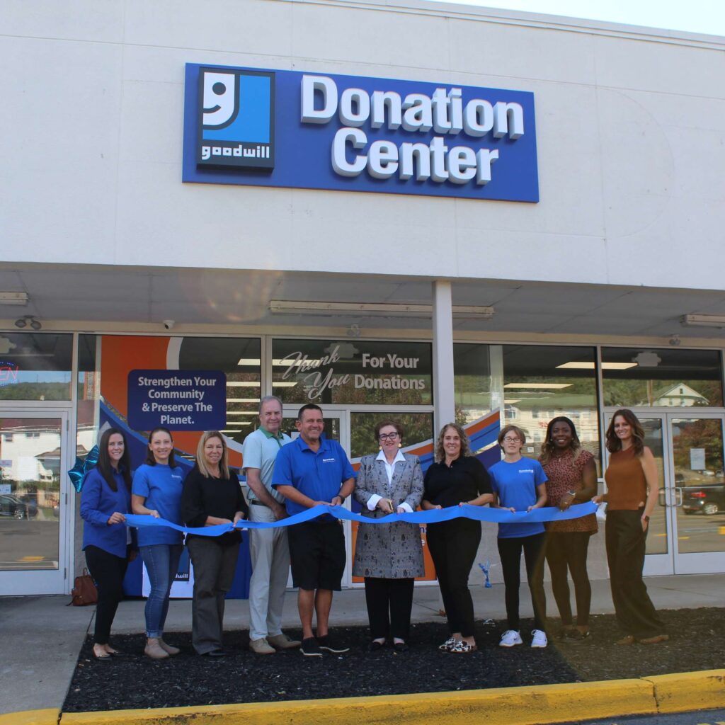A group of individuals gather in front of a Goodwill Attended Donation Center and cut a ribbon as part of a grand opening celebration